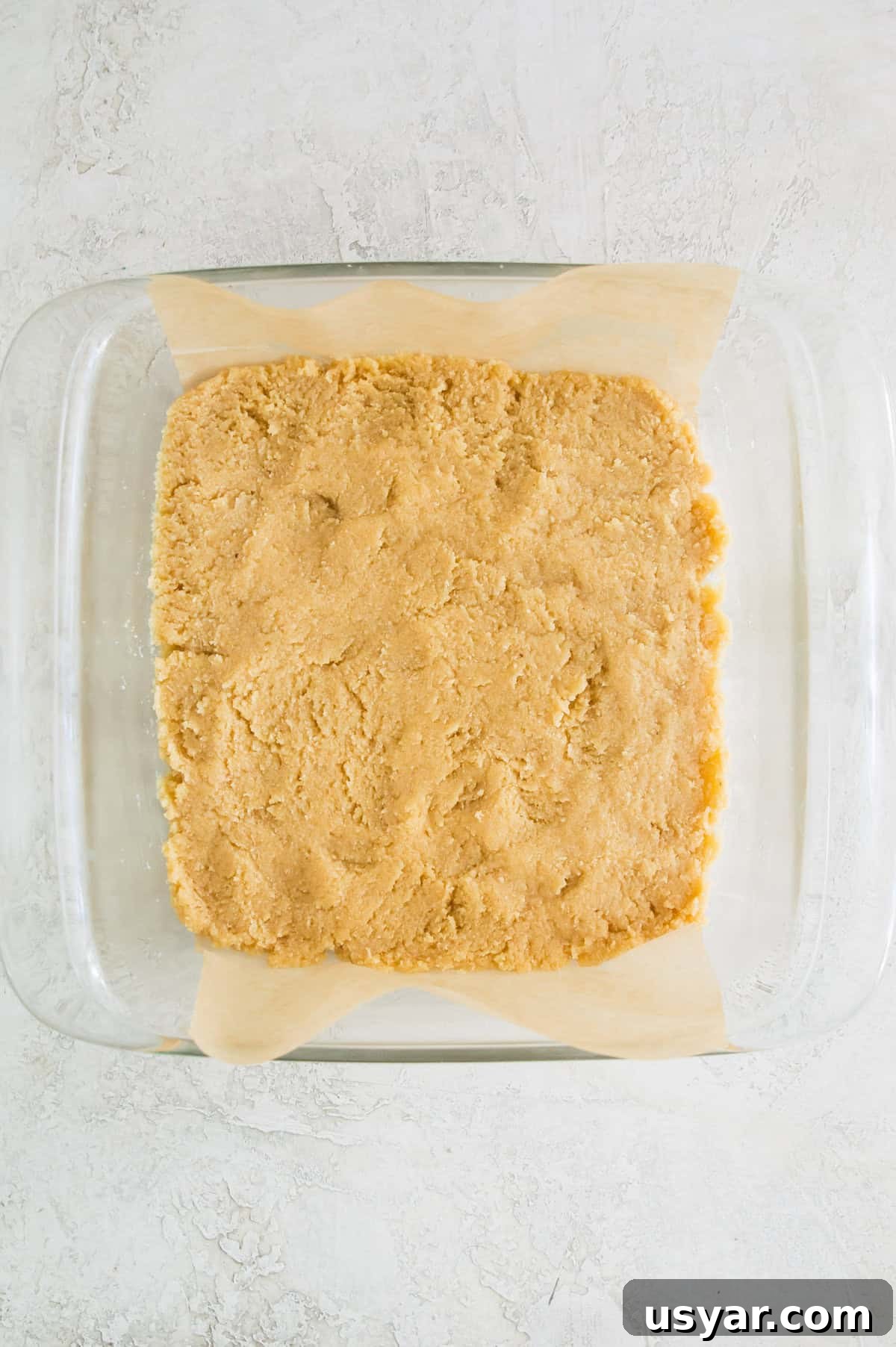 Gluten-Free Vegan and Paleo Apple Pie Bars 5 A square baking pan lined with parchment paper with a shortbread dough in it.