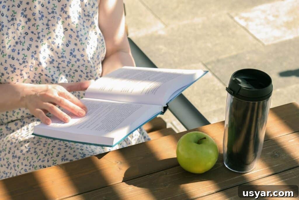 woman reading book on her lunch break at a city table with a reusable coffee cup and apple