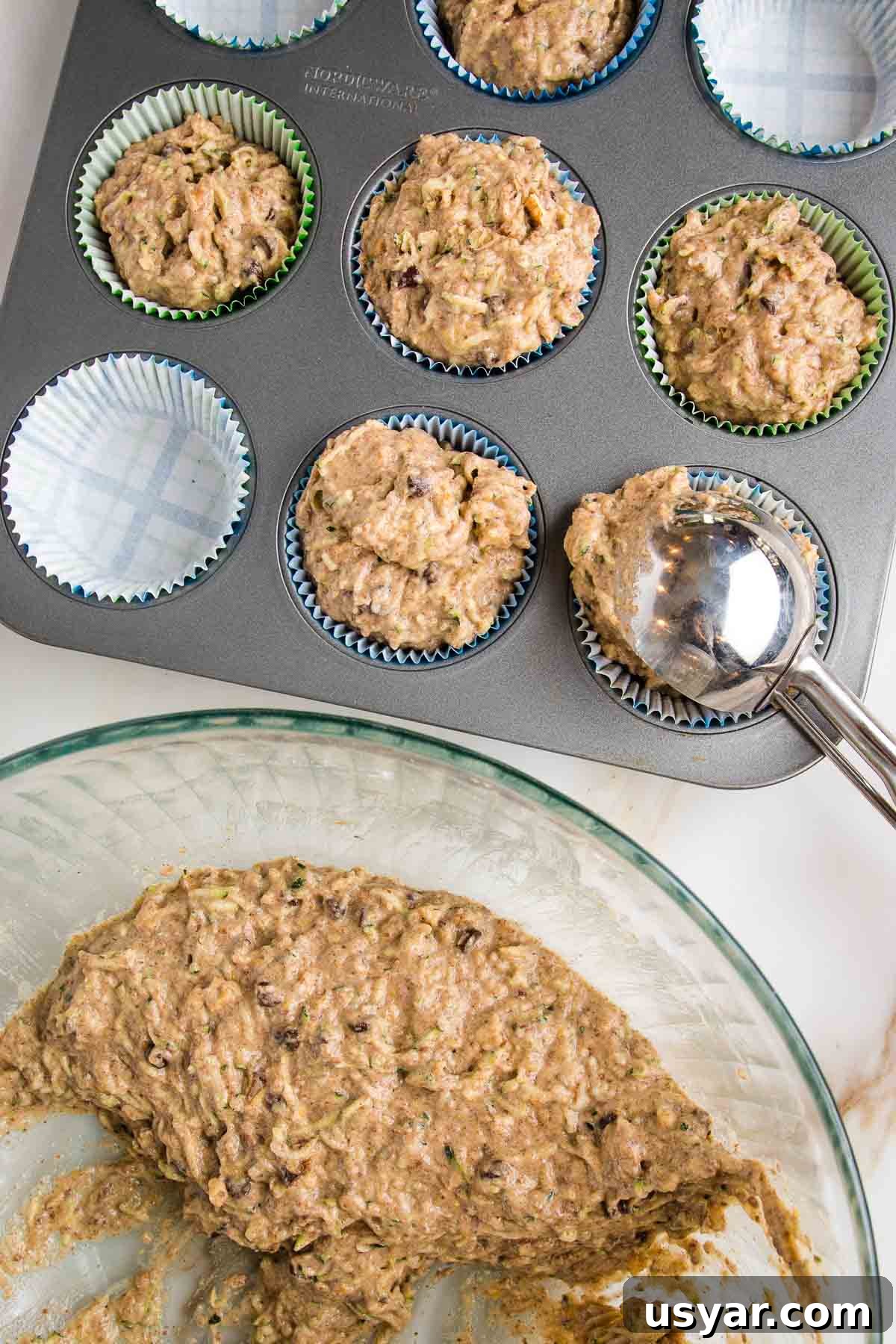 Zucchini muffin batter in a large mixing bowl.  Cookie scoop portioning batter into muffin tin lined with parchment paper liners.