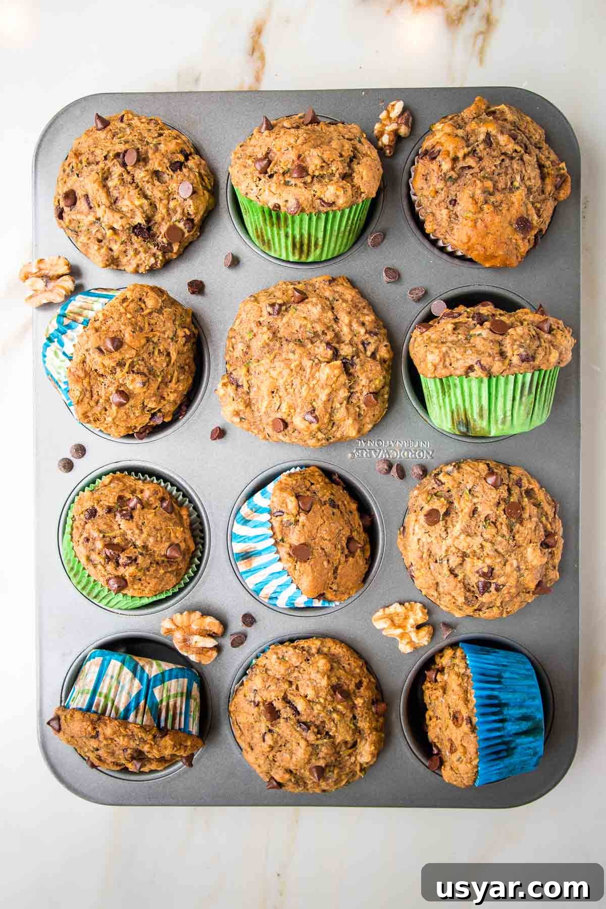 Chocolate Chip Zucchini Muffins in a muffin tin. Some are turned on their side and there are mini chocolate chips and walnuts strewn about.
