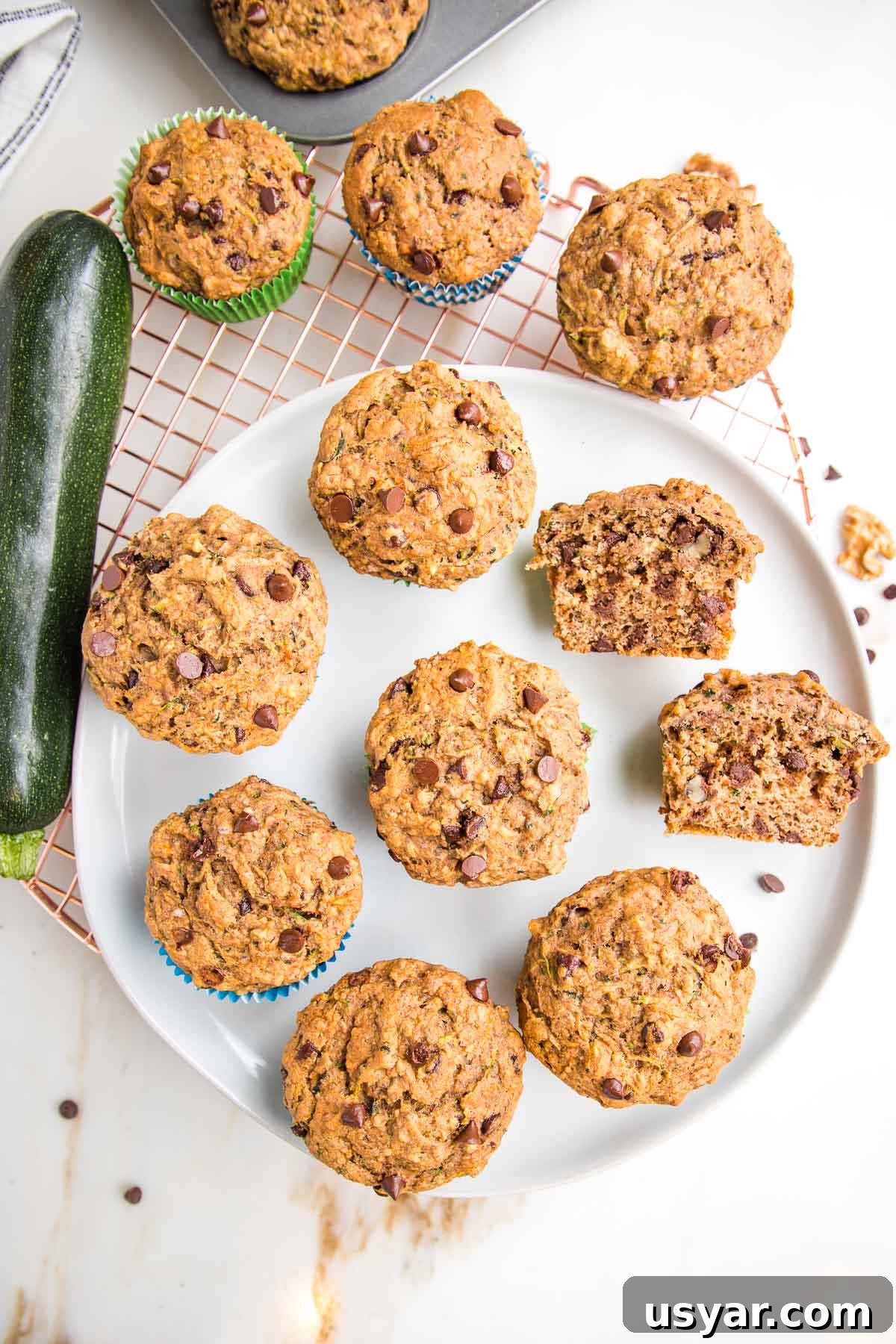 Plate of zucchini muffins, one is cut open and the texture is uneven, the chocolate chips melty. There are a few more funnis on a wire rack and a whole zucchini off to the side.