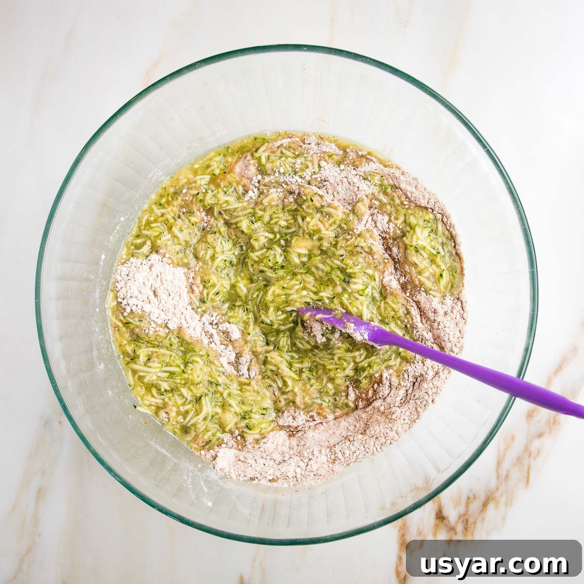 Large glass mixing bowl with shredded zucchini mixture and a purple spatula mixing that together with the dry ingredients.