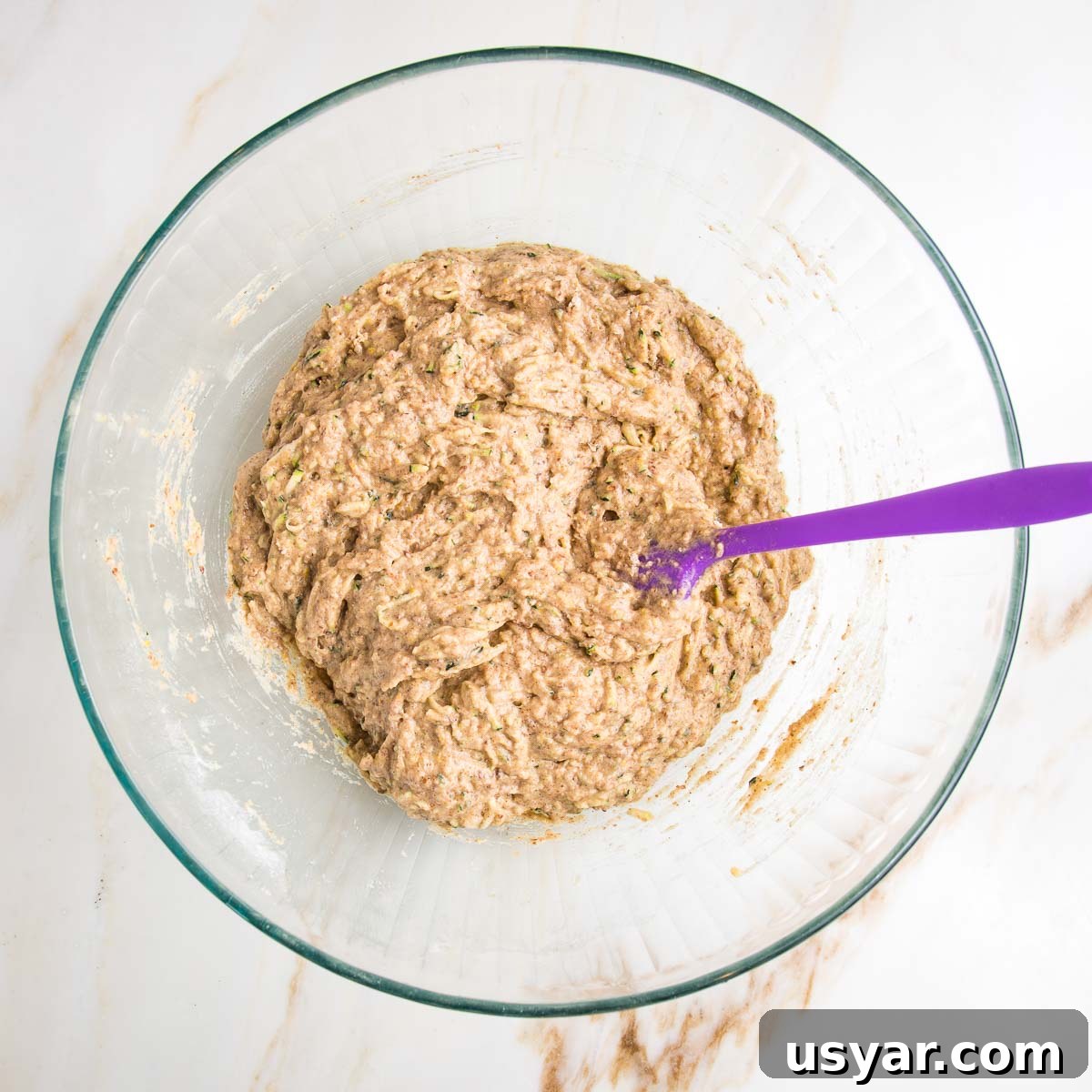 Large glass mixing bowl with lumpy zucchini muffin batter and a purple spatula.