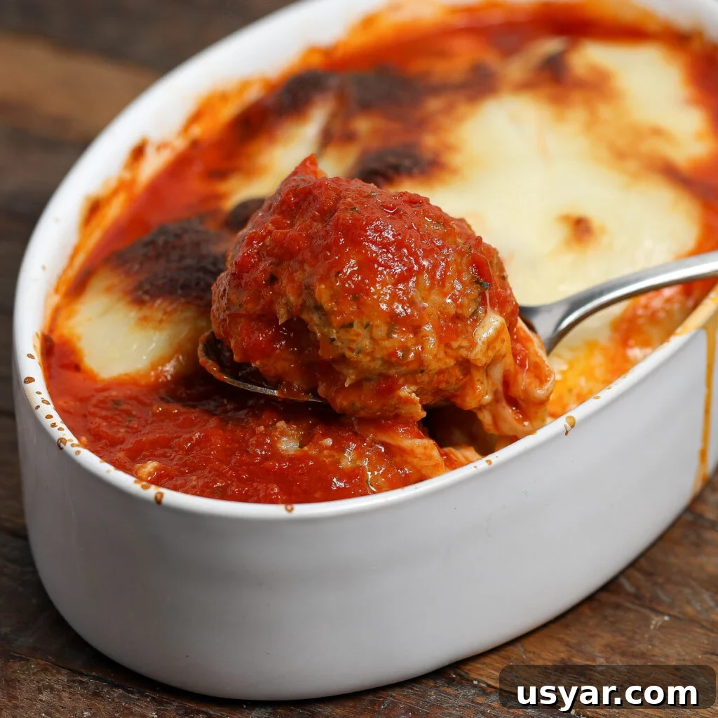 A close-up of a Chicken Parmesan Meatball covered in tomato sauce being lifted from a white ceramic dish filled with saucy, cheesy baked meatballs. The dish rests on a wooden surface.