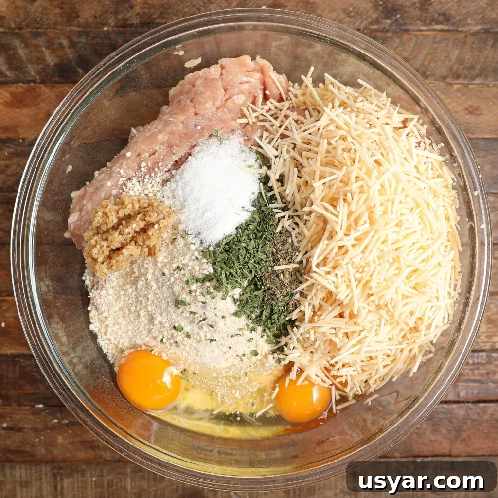 A glass bowl on a wooden surface contains all the ingredients for Chicken Parmesan Meatballs—ground meat, shredded cheese, breadcrumbs, eggs, minced garlic, dried herbs, and salt—each neatly arranged in separate piles.