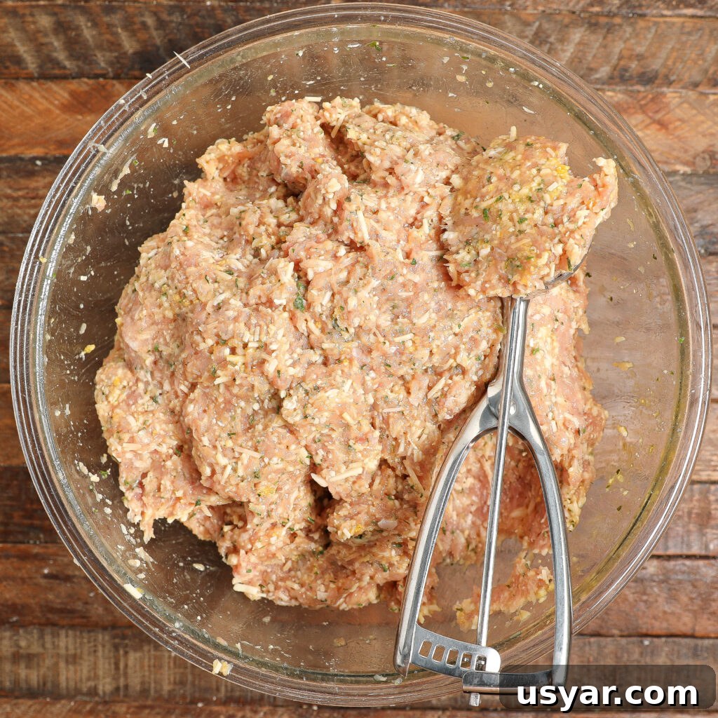A glass bowl filled with a mixture for Chicken Parmesan Meatballs—ground meat, herbs, and spices—sits on a wooden surface. A metal scoop rests inside the bowl, partially submerged in the savory blend.