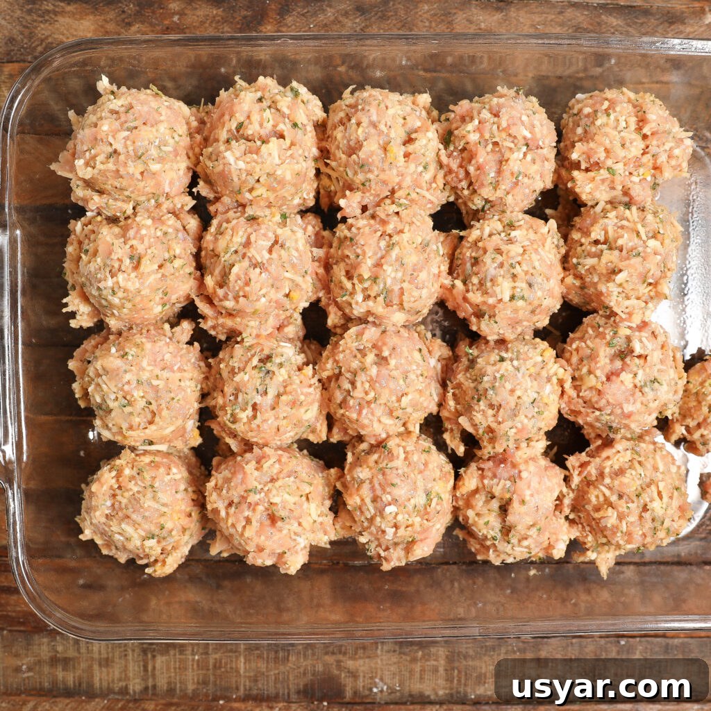 A glass baking dish filled with evenly spaced, uncooked Chicken Parmesan Meatballs made from a mixture of ground meat and herbs, arranged in neat rows on a wooden surface.