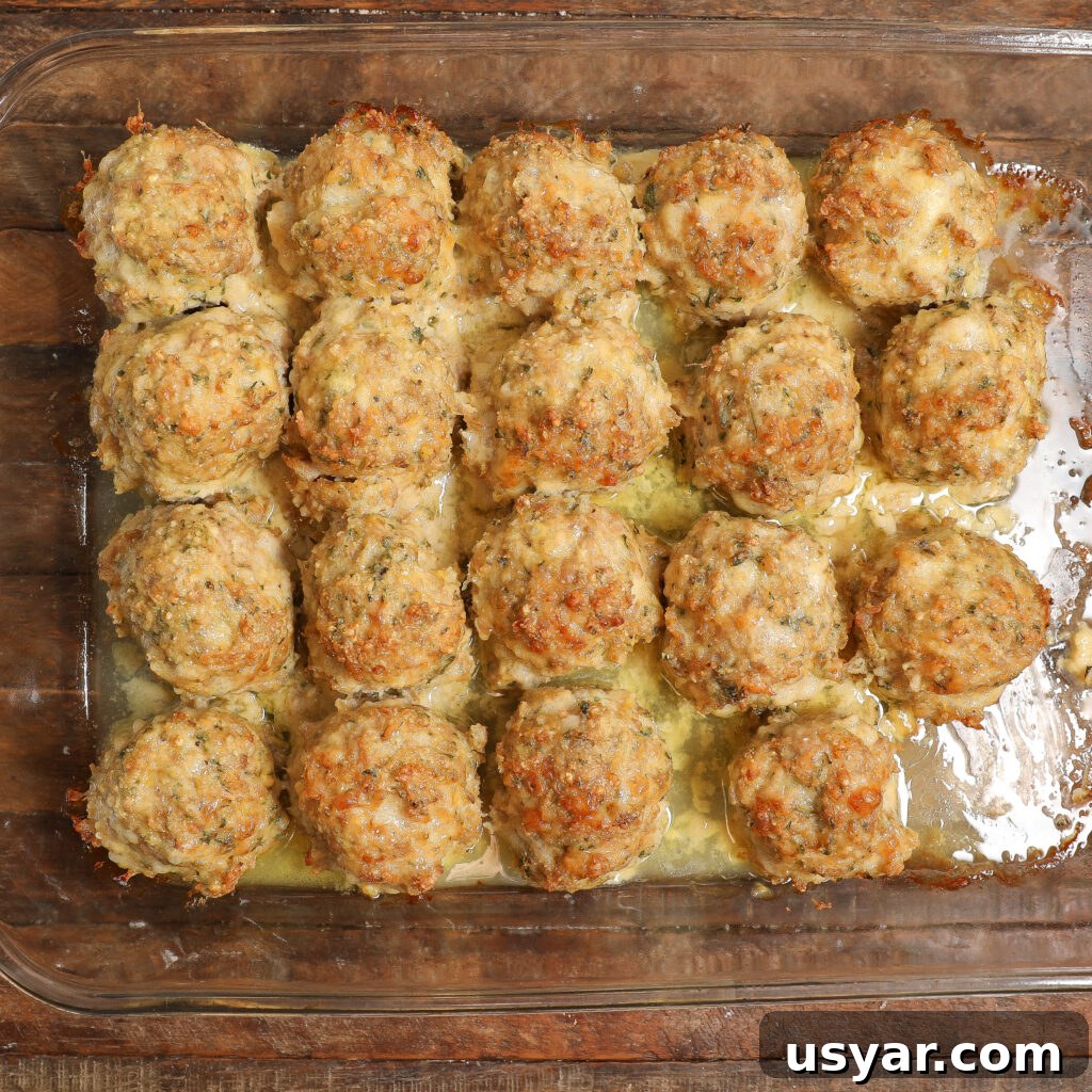 A glass baking dish filled with twenty golden-brown Chicken Parmesan Meatballs, arranged in a 4x5 grid, rests in a layer of melted cheese and savory juices on a wooden surface.