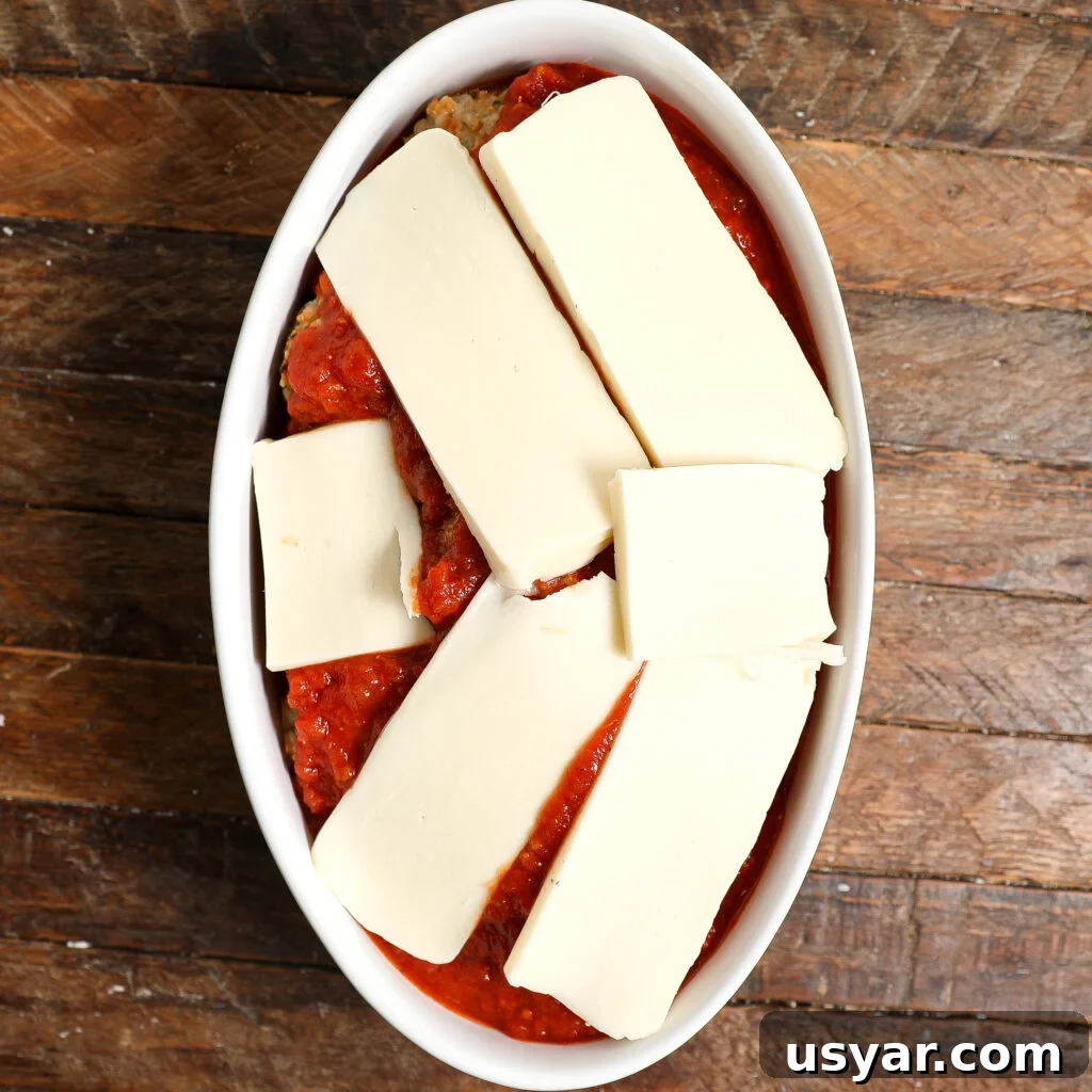 An oval baking dish filled with tomato sauce and Chicken Parmesan Meatballs, topped with several thick rectangular slices of mozzarella cheese, placed on a wooden surface.