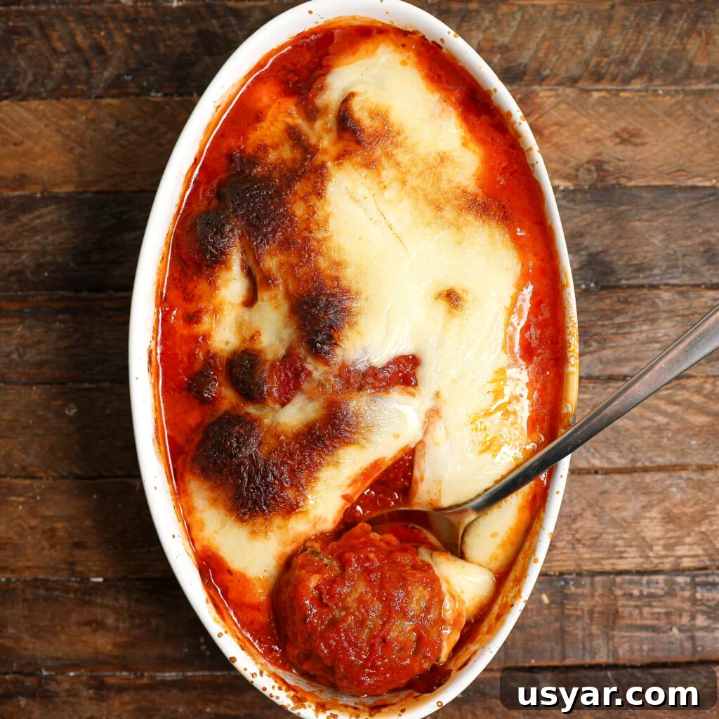 Oval dish filled with baked cheesy lasagna in red tomato sauce, topped with golden Chicken Parmesan Meatballs and slightly browned on top, with a serving spoon scooping out a portion; dish sits on a rustic wooden table.