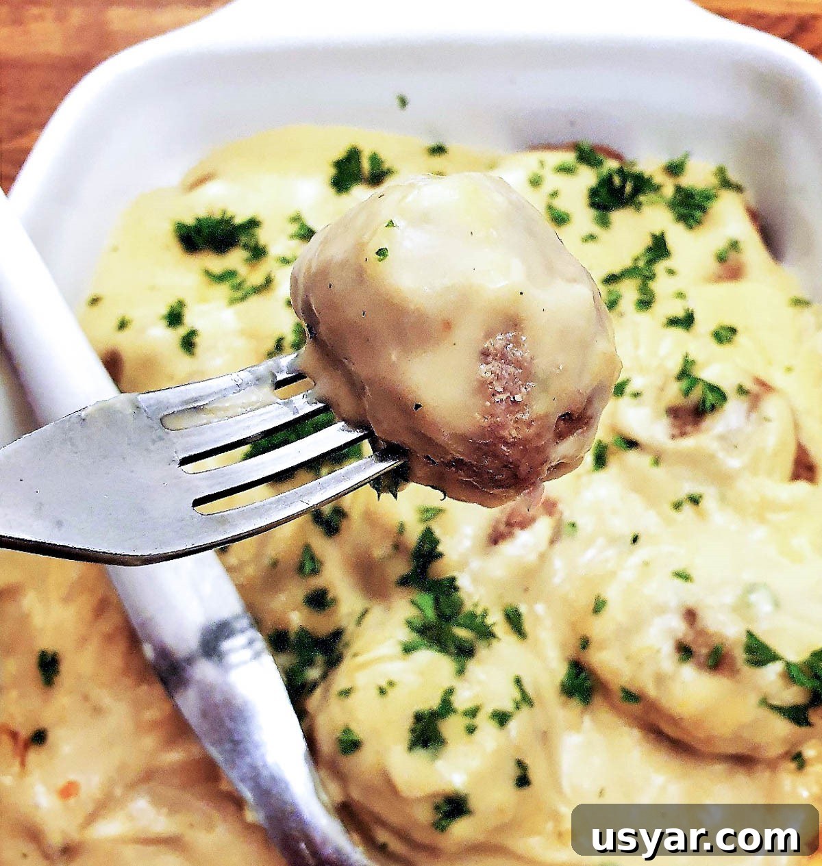 A Swedish meatball on a fork, above a dish of meatballs covered with creamy mustard sauce.