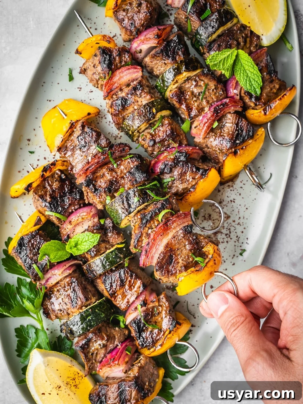 Authentic Middle Eastern Beef Shish Kebab Recipe with Sides 2 Six grilled Middle Eastern Beef Shish Kebabs with yellow bell peppers, zucchini, and red onions on skewers, garnished with fresh herbs and served with lemon wedges on a white platter. A hand is holding one skewer.