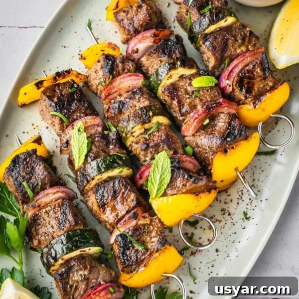 Middle Eastern Beef Shish Kebabs with yellow bell peppers, red onions, and zucchini on skewers, garnished with fresh mint and parsley. Served on a white platter with a lemon wedge and a small bowl of sauce.