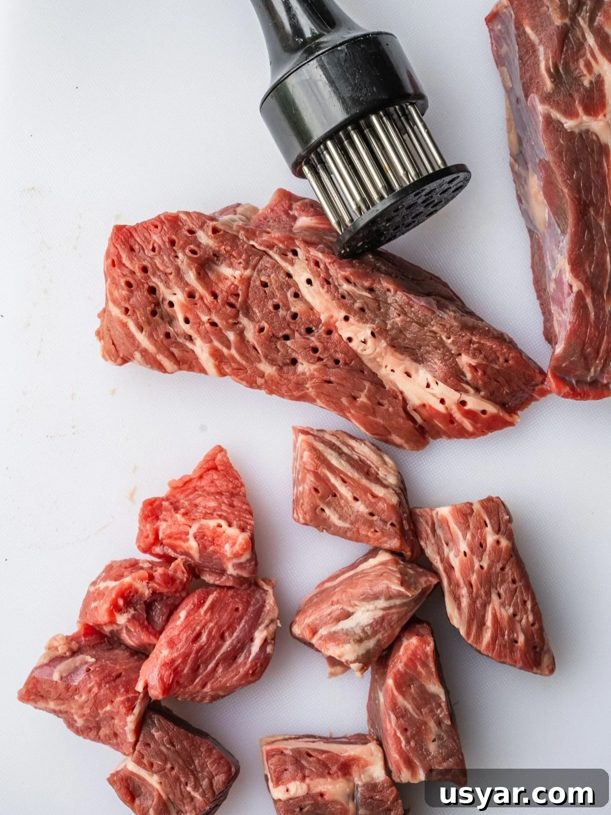 Authentic Middle Eastern Beef Shish Kebab Recipe with Sides 4 Raw beef pieces on a white cutting board, some pierced with holes from a black meat tenderizer tool resting above them.