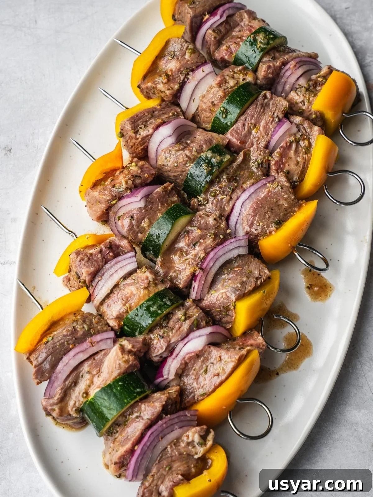 Authentic Middle Eastern Beef Shish Kebab Recipe with Sides 7 Six metal skewers with marinated Middle Eastern Beef Shish Kebabs with beef cubes, yellow bell peppers, red onion slices, and zucchini on an oval plate.