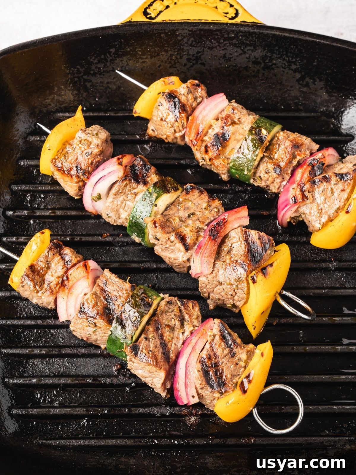 Authentic Middle Eastern Beef Shish Kebab Recipe with Sides 8 Three grilled beef kabobs with yellow bell peppers, red onion, and green zucchini on metal skewers are cooking on a black grill pan with visible grill marks.