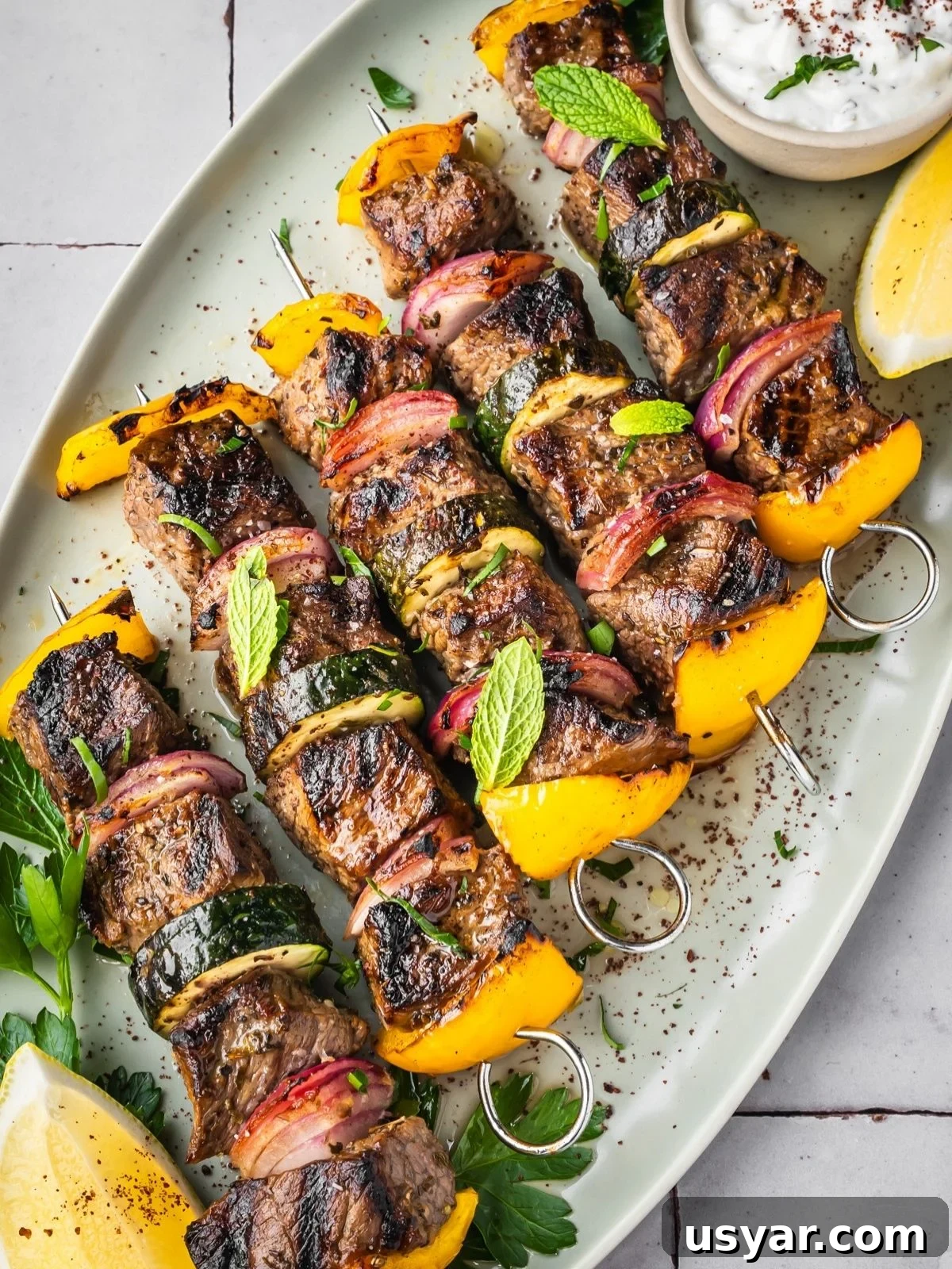 Authentic Middle Eastern Beef Shish Kebab Recipe with Sides 9 Middle Eastern Beef Shish Kebabs with yellow bell peppers, red onions, and zucchini on skewers, garnished with fresh mint and parsley. Served on a white platter with a lemon wedge and a small bowl of sauce.