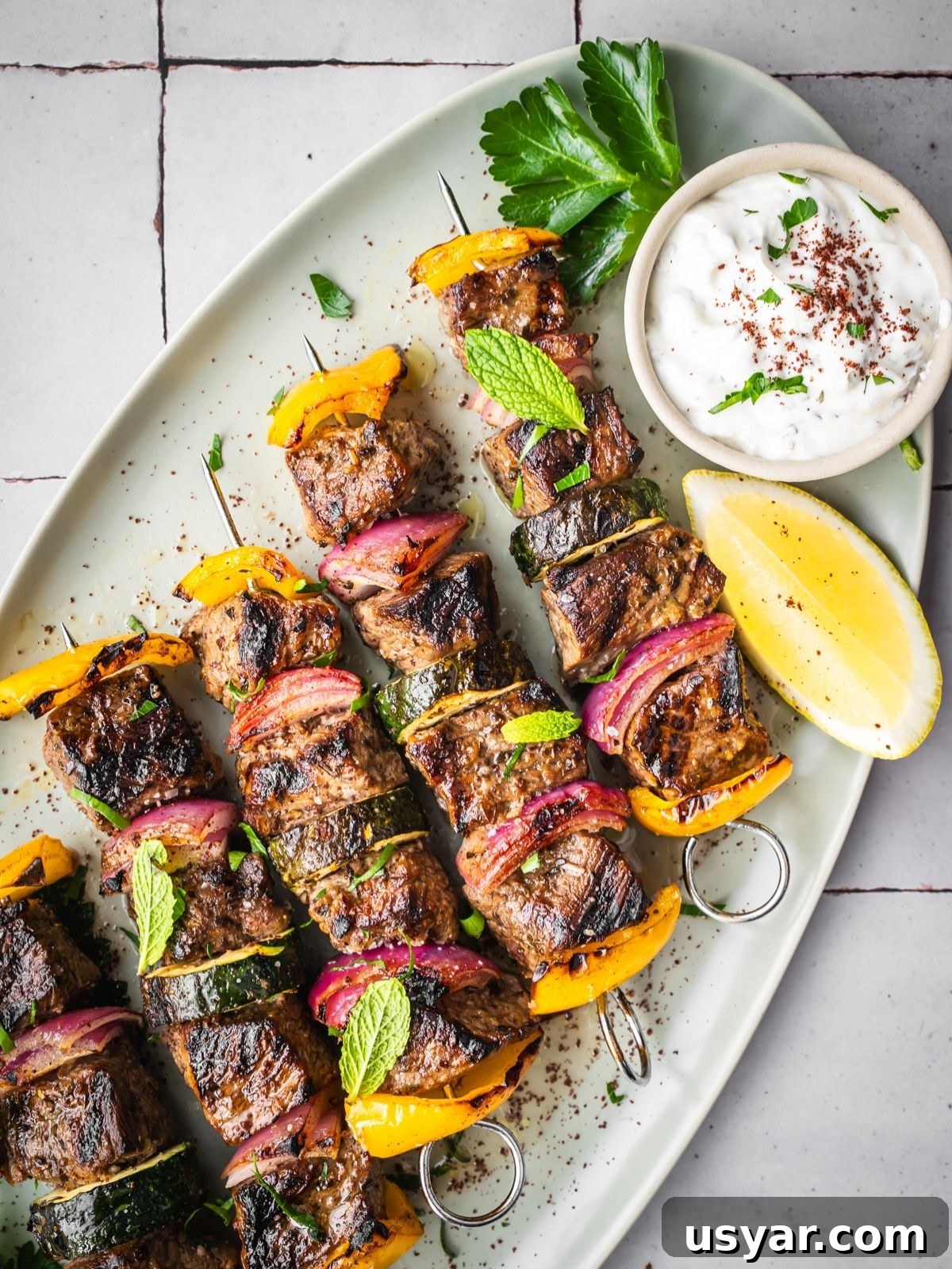 Authentic Middle Eastern Beef Shish Kebab Recipe with Sides 10 Middle Eastern Beef Shish Kebabs with yellow bell peppers, red onions, and zucchini on skewers, garnished with fresh mint and parsley. Served on a white platter with a lemon wedge and a small bowl of sauce.