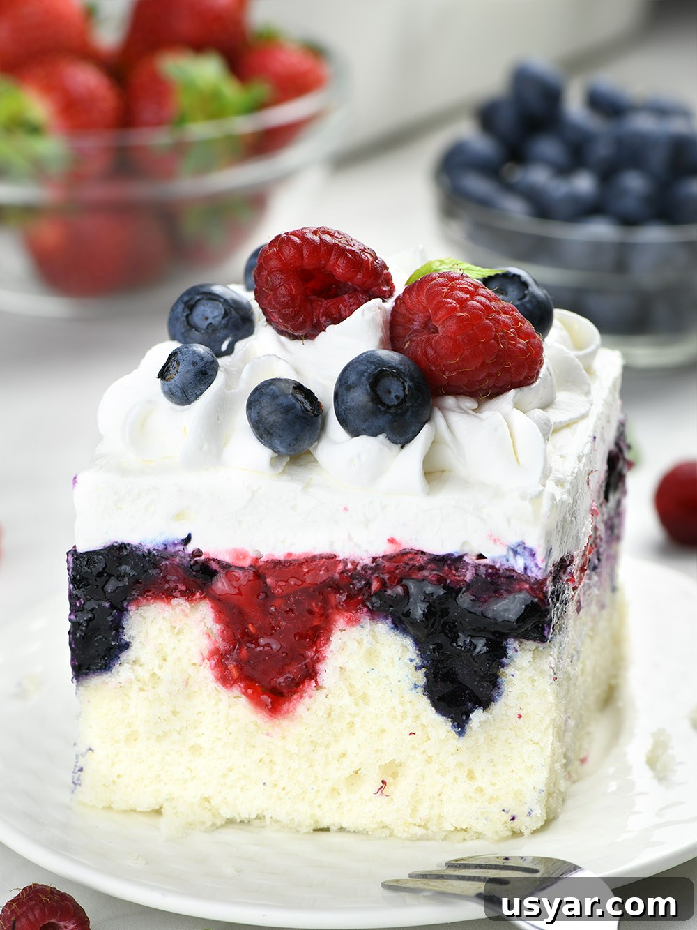 Summer Berry Poke Cake Recipe with Fresh Berries 9 Summer berry poke cake on a white plate, garnished with berries and topped with cool whip.
