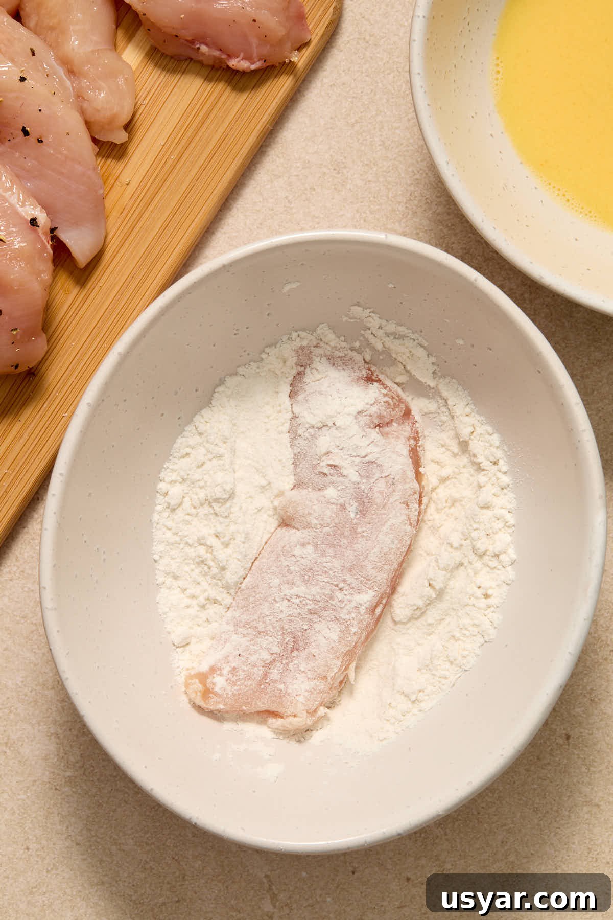 A chicken strip being dredged in flour in a white bowl.