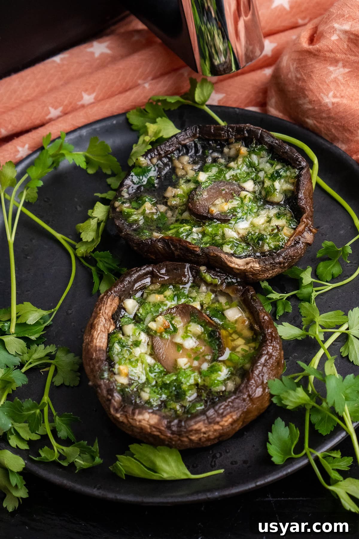 Two cooked Portobello mushrooms with melted garlic butter inside the mushroom served on a black plate with fresh parsley around them and an air fryer in the background