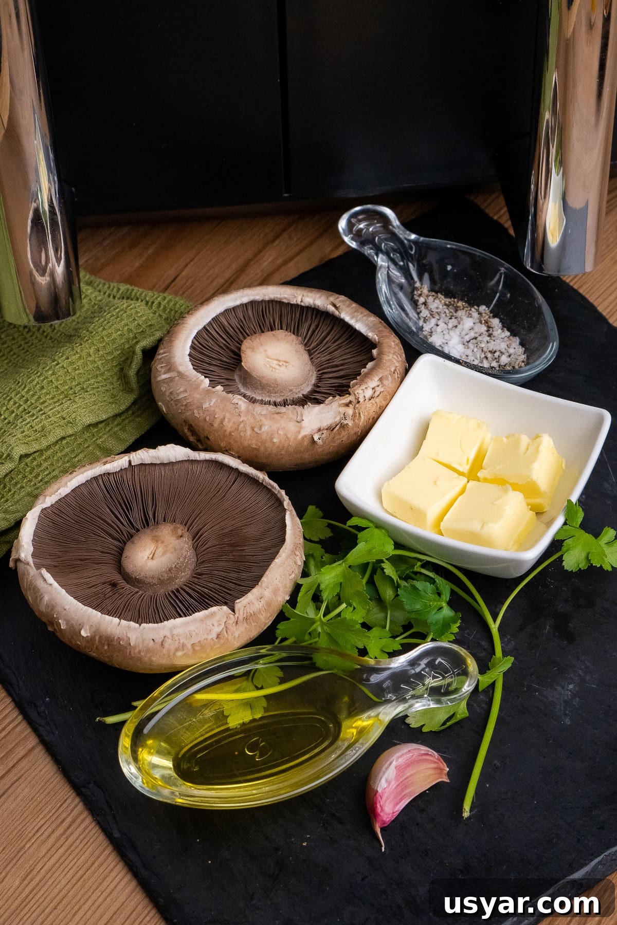 Two Portobello mushrooms, olive oil, cubes of butter, a garlic clove, salt, pepper and fresh parsley on a slate board with an air fryer in the background