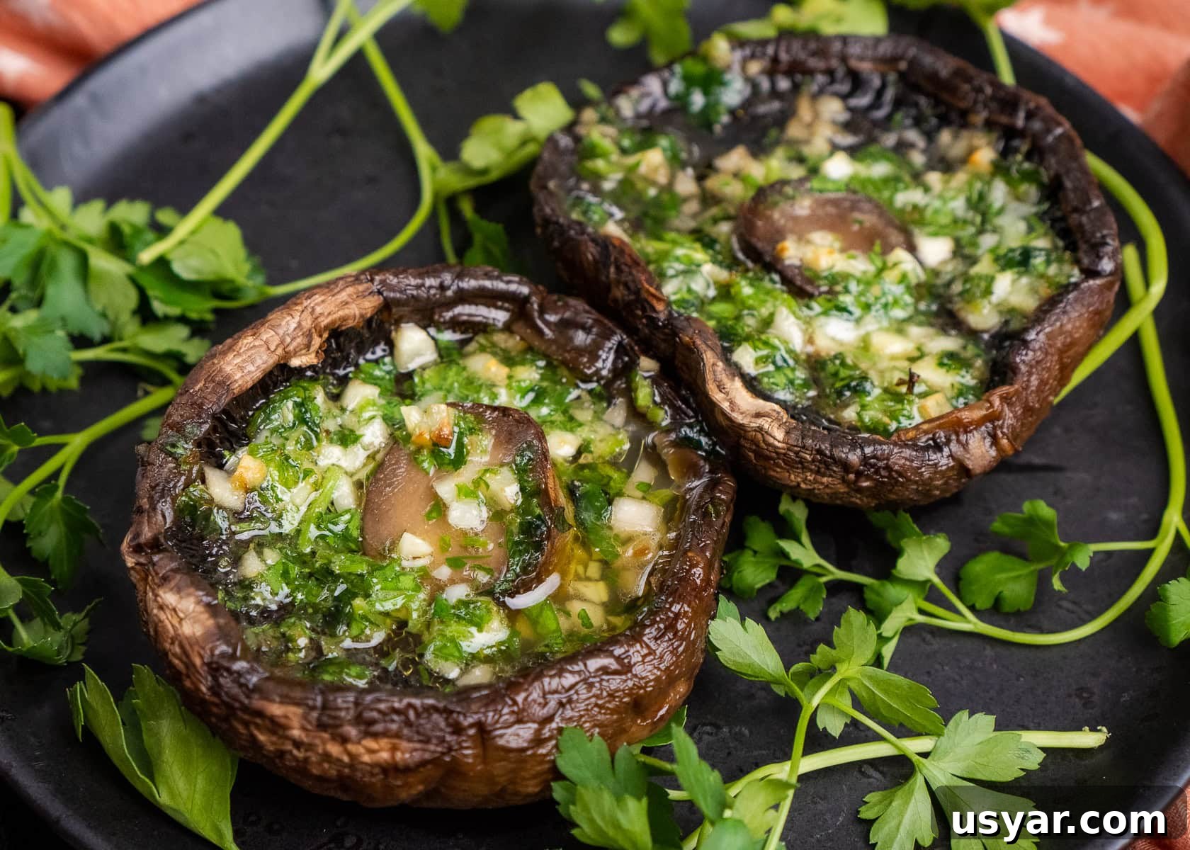 Two cooked Portobello mushrooms with melted garlic butter inside the mushroom served on a black plate with fresh parsley around them