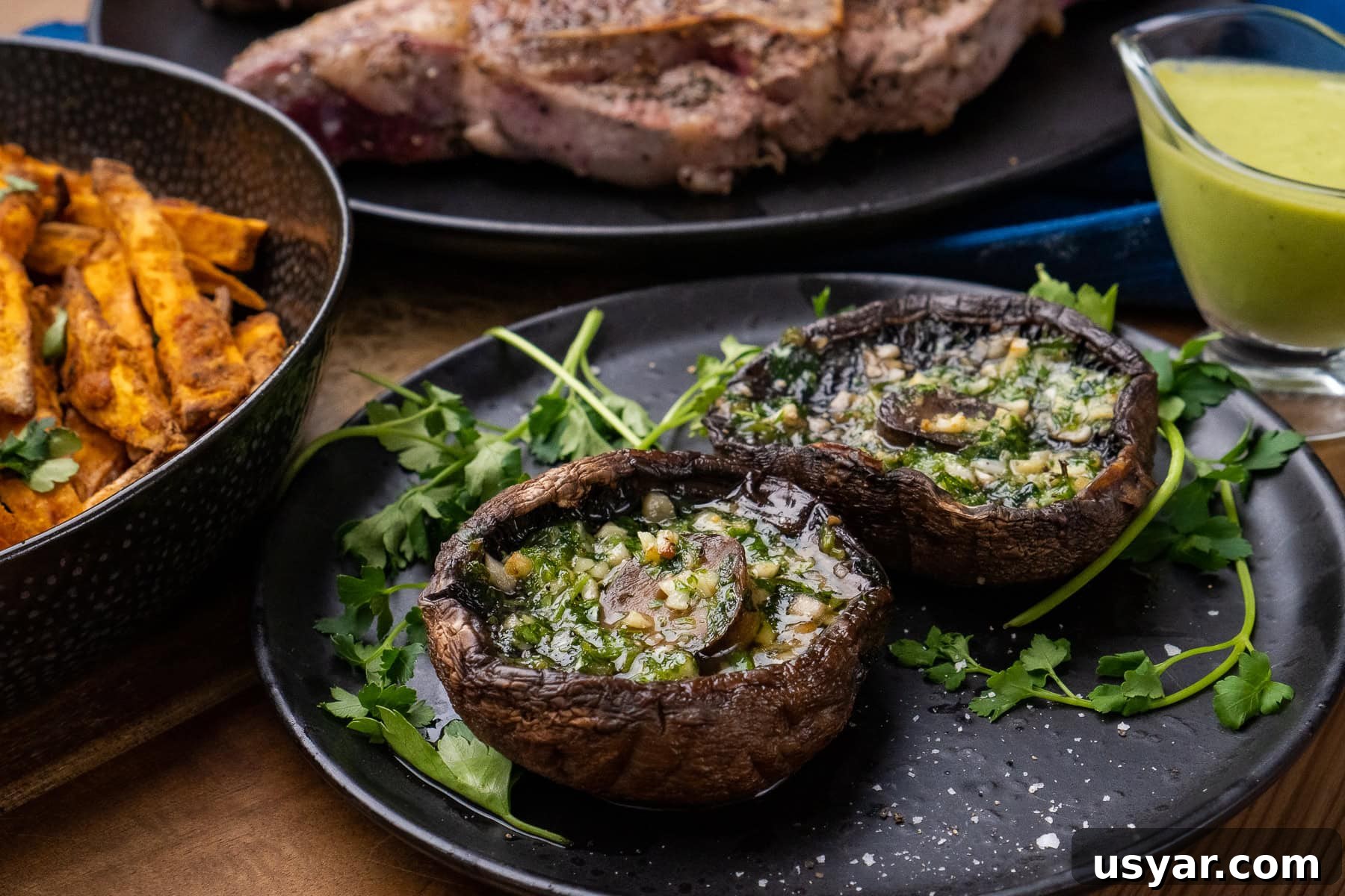 Two cooked Portobello mushrooms with melted garlic butter inside the mushroom served on a black plate with fresh parsley around them and sea salt sprinkled over the top and in the background is Barnsley chops, sweet potato fries and minted pea puree