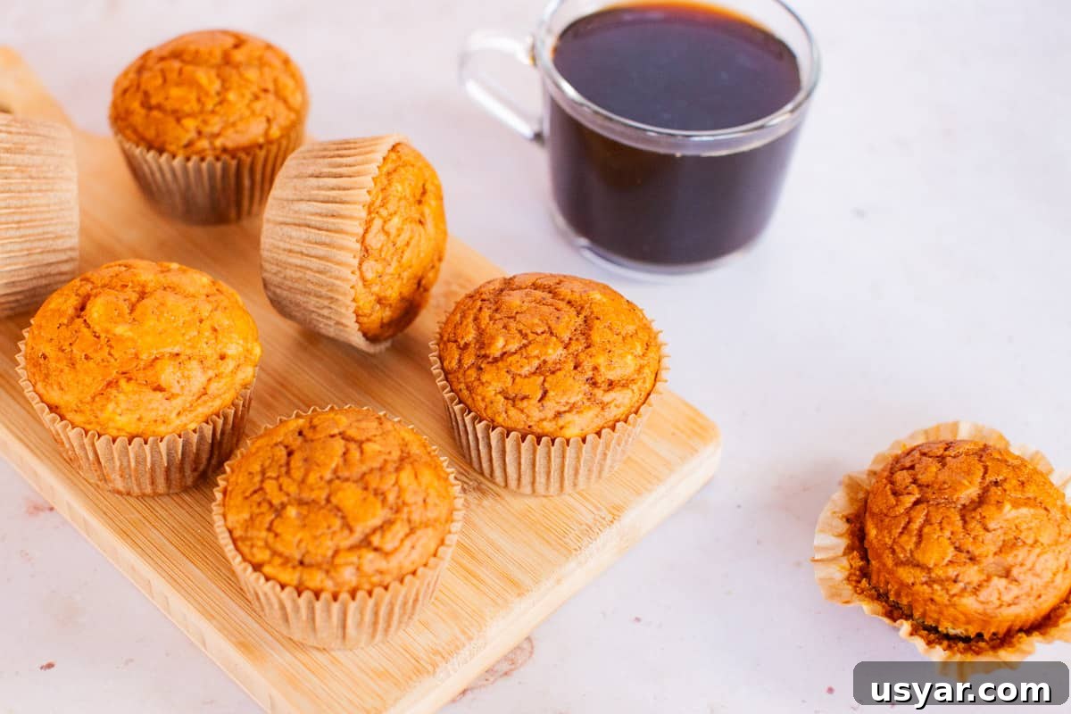 Moist Pumpkin Spice Muffins Recipe 4 Pumpkin muffins on a wooden serving dish arranged on a marble countertop with a clear, glass mug filled with coffee.