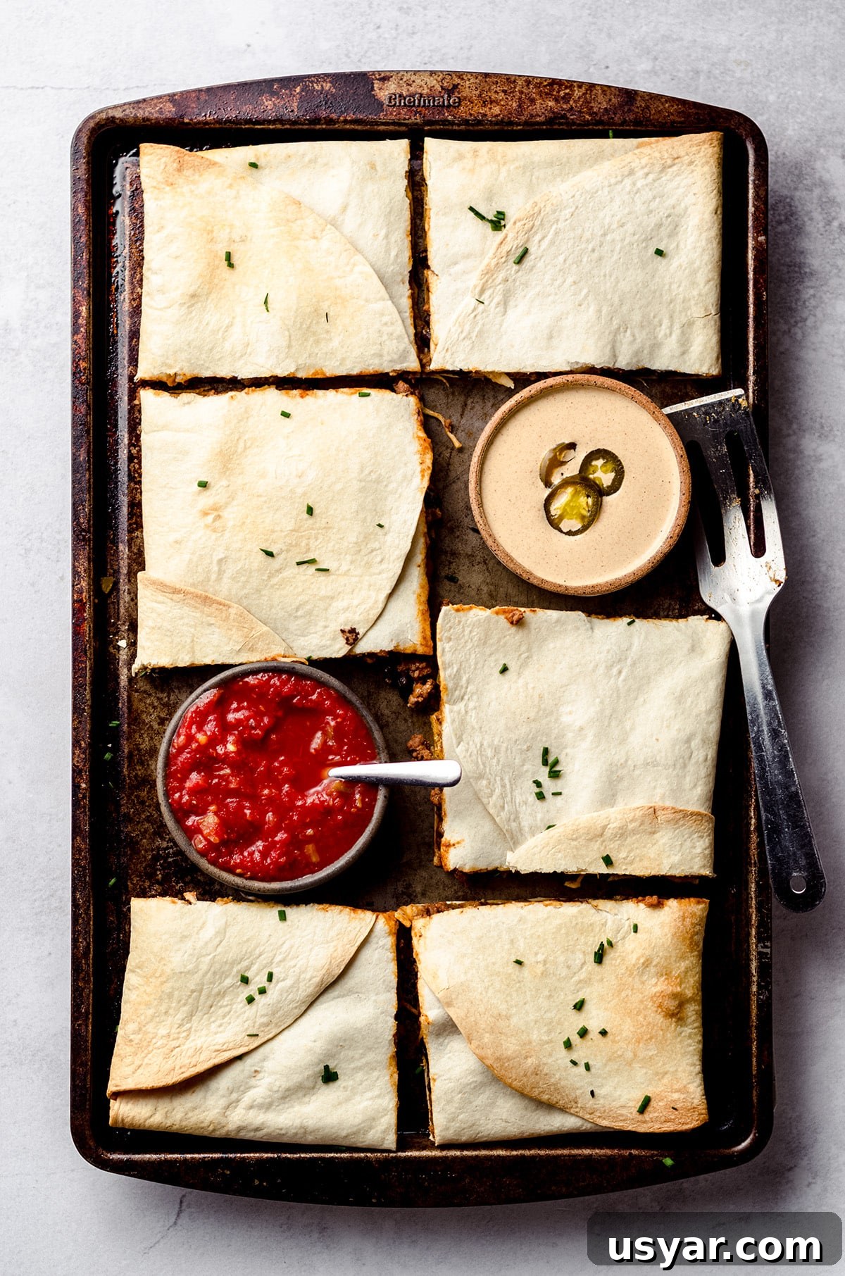 An overhead shot of a sheet pan filled with square quesadillas and served with bowls of sauce and salsa.