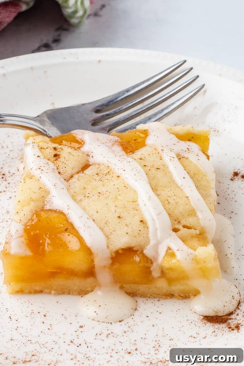 Cinnamon-Apple Pie Bars with Vanilla Glaze 9 Close up image of apple pie bars on white plate with fork.
