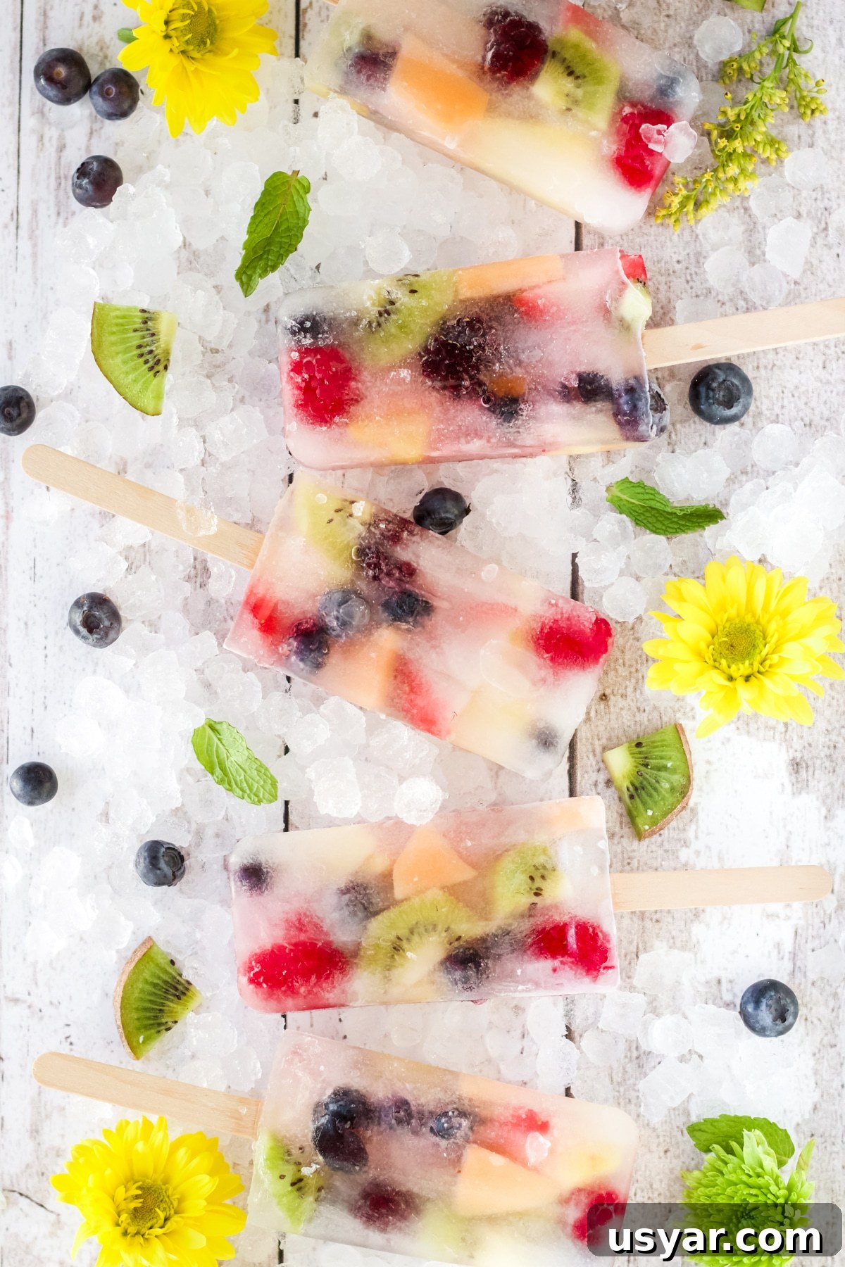5 fruit popsicles laying on crushed ice board decorated with yellow flowers and fruit