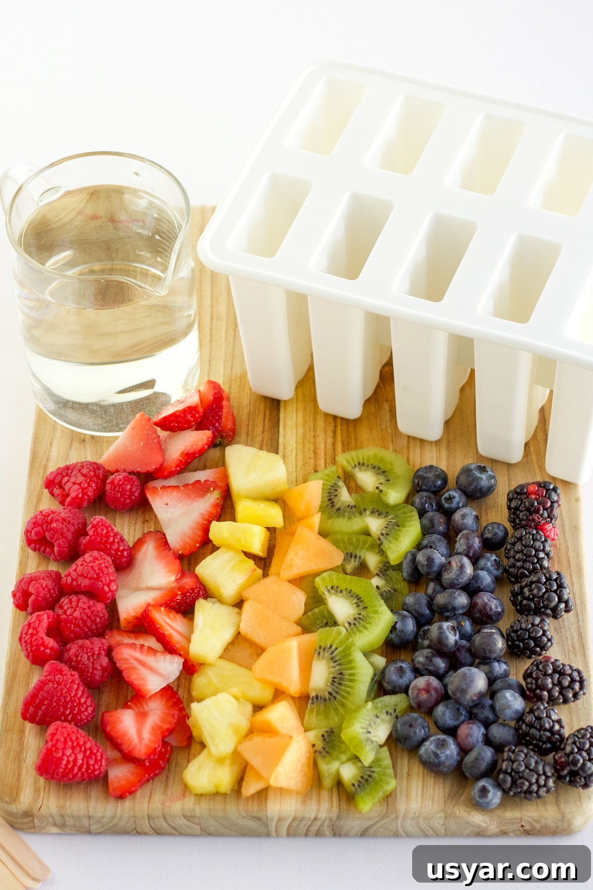rainbow colored cut fruit on a cutting board with a glass of clear juice and a popsicle mold