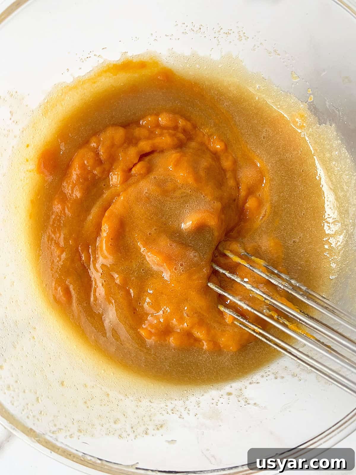 Mixing pumpkin puree into oil and sugar batter in a glass bowl with a wire whisk.