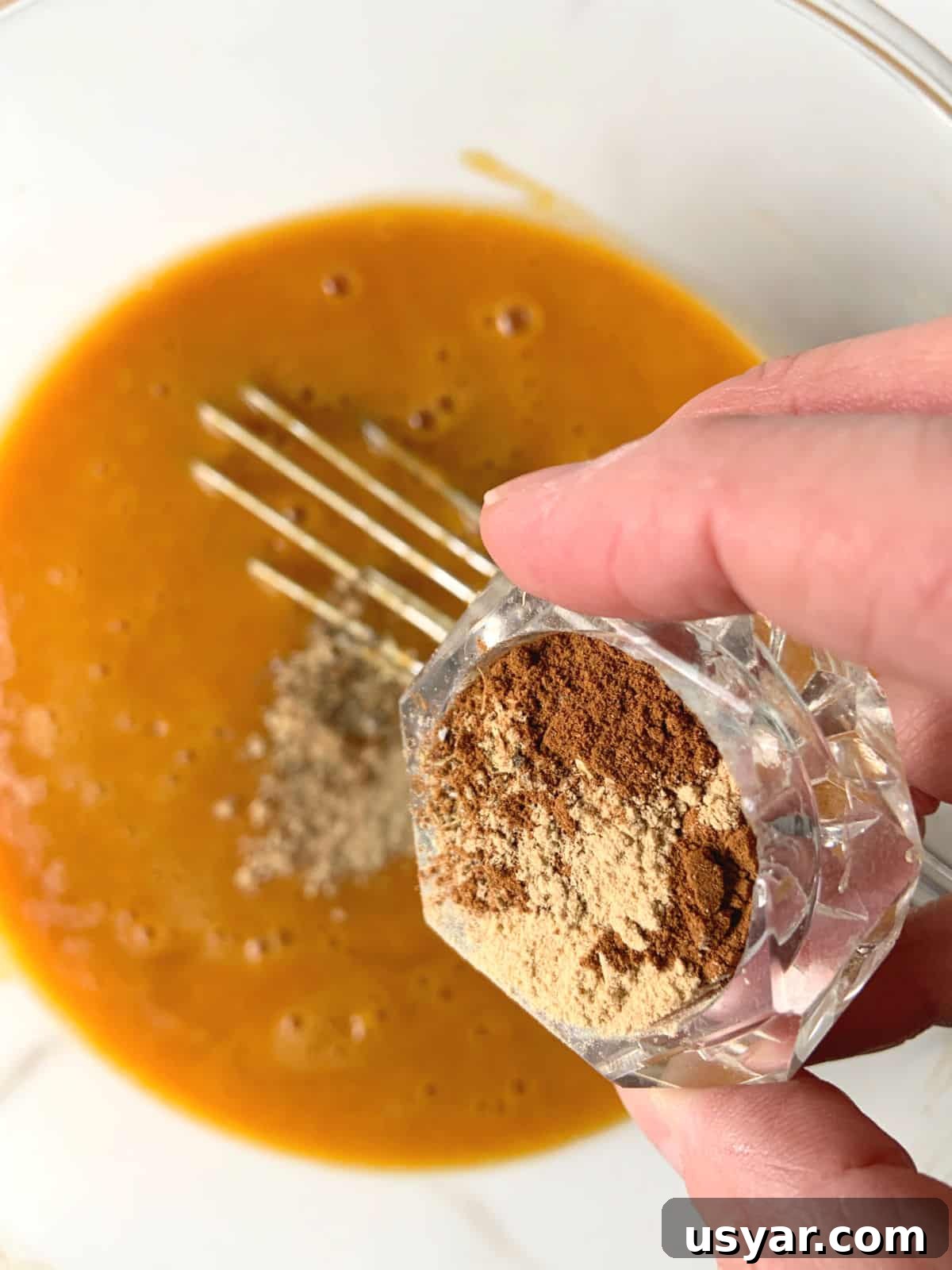 Adding pumpkin pie spice mix to pumpkin batter in a glass bowl. 
