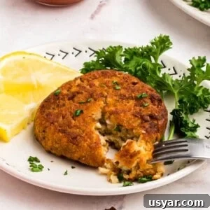 Golden crispy crab cake on a small white plate with a fork removing a bite and with a small bowl of cocktail sauce on the table for dipping.