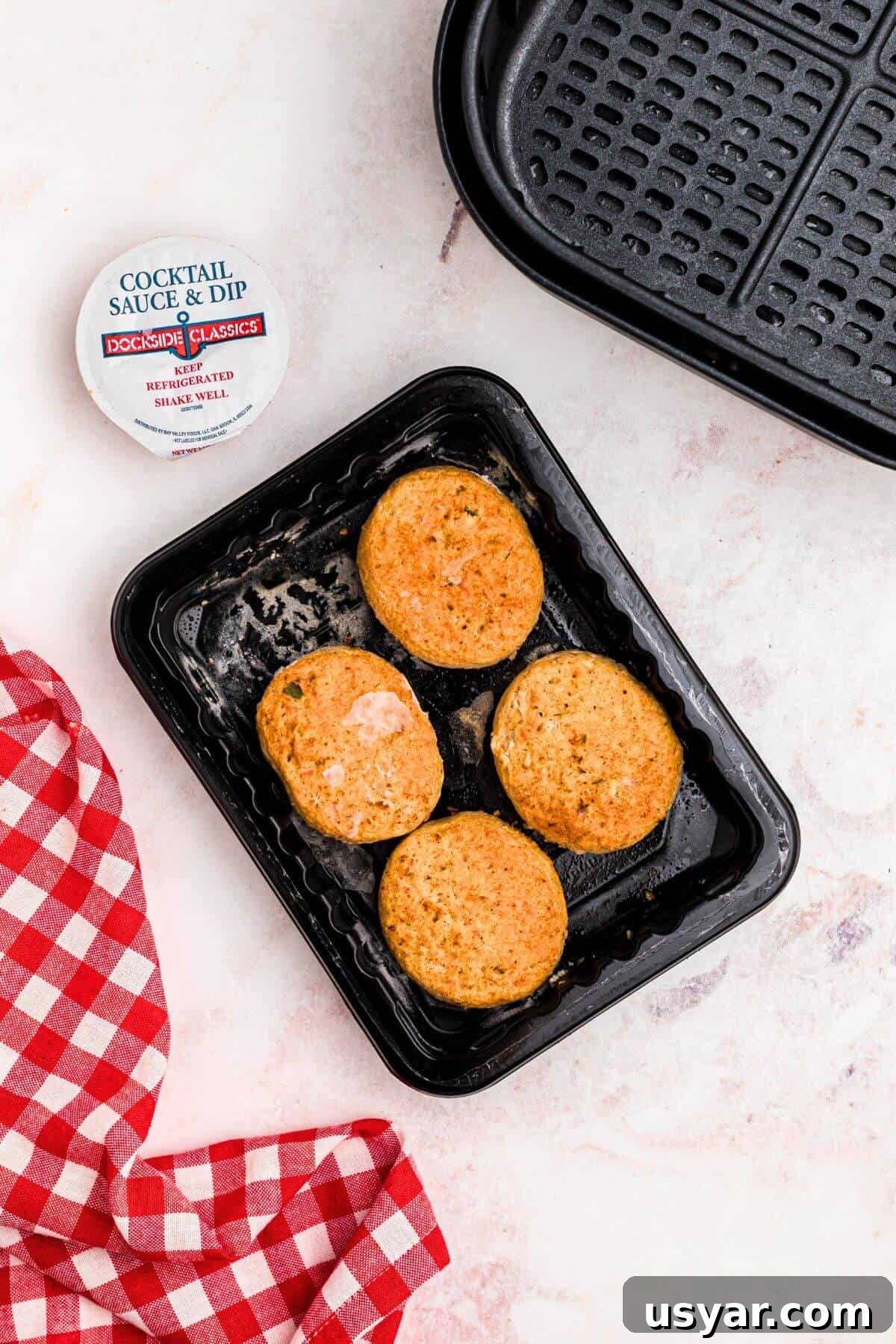 Frozen crab cakes on a black plastic plate in front of the air fryer basket. 
