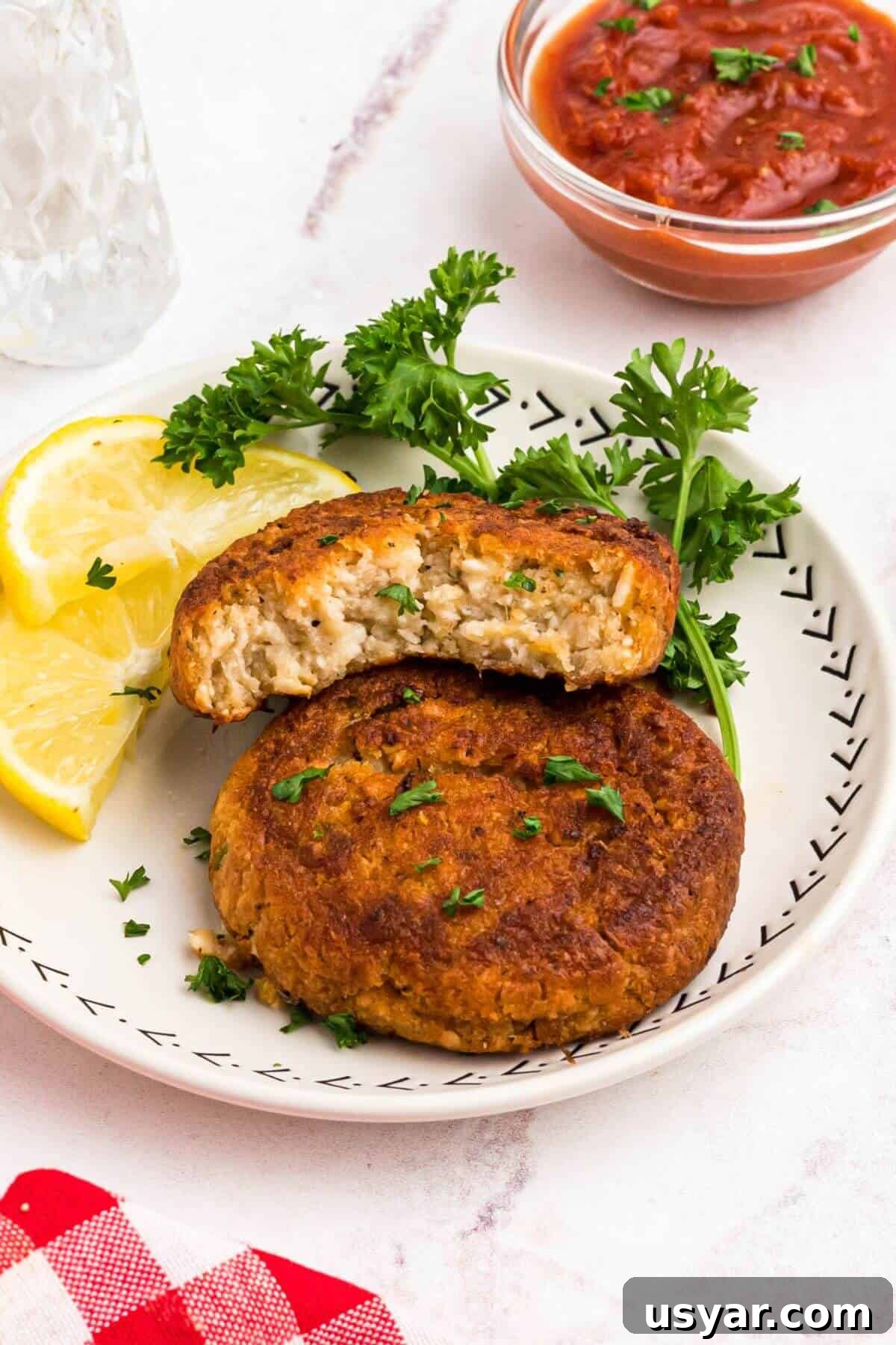 Golden crispy crab cakes on a small white plate with a small bowl of cocktail sauce on the table for dipping. 