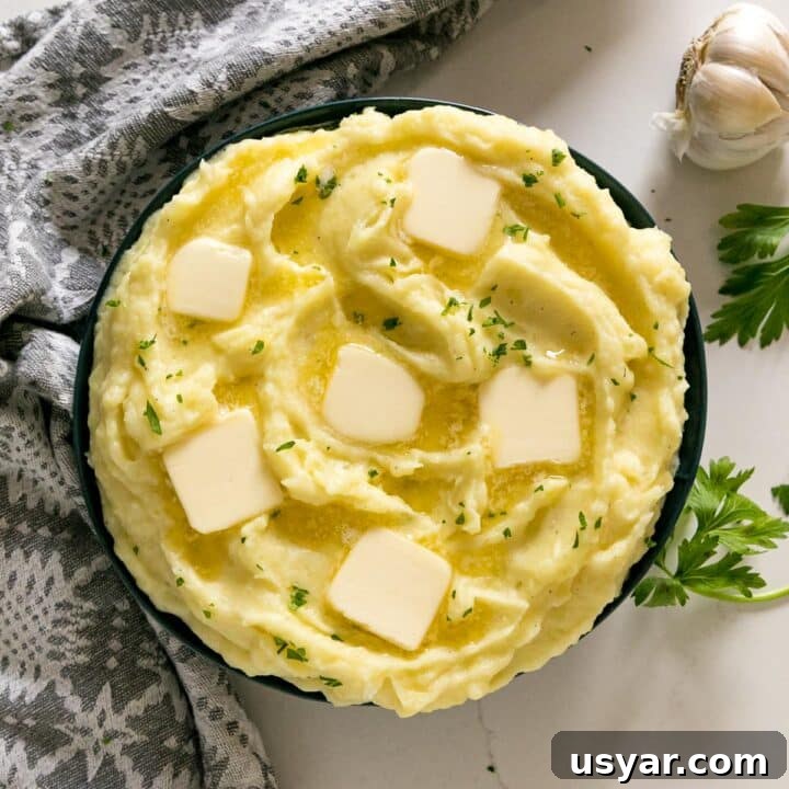 An overhead image of a bowl of mashed potatoes with melting pats of butter and chopped parsley on top with a head of garlic, grey napkin and sprigs of parsley round it.