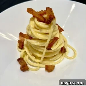 Secret Tips for Authentic Carbonara Pasta 3 A plate of spaghetti carbonara is neatly presented in a swirl topped with crispy bacon pieces. The creamy pasta is lightly coated in sauce and garnished with black pepper, served on a white dish.