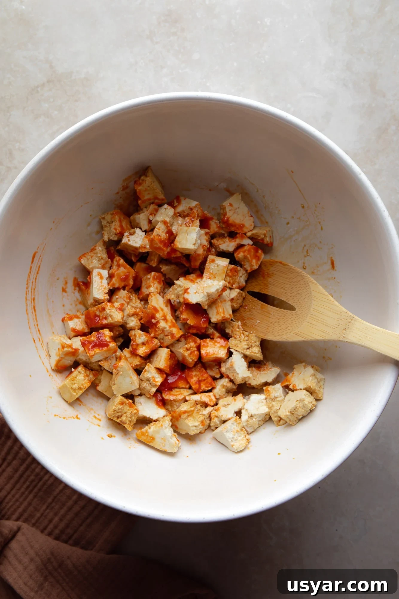 Vegan Banh Mi Sandwich with Pickled Vegetables 4 using a wooden spoon to toss tofu cubes in hot sauce and seasonings in a large white bowl.