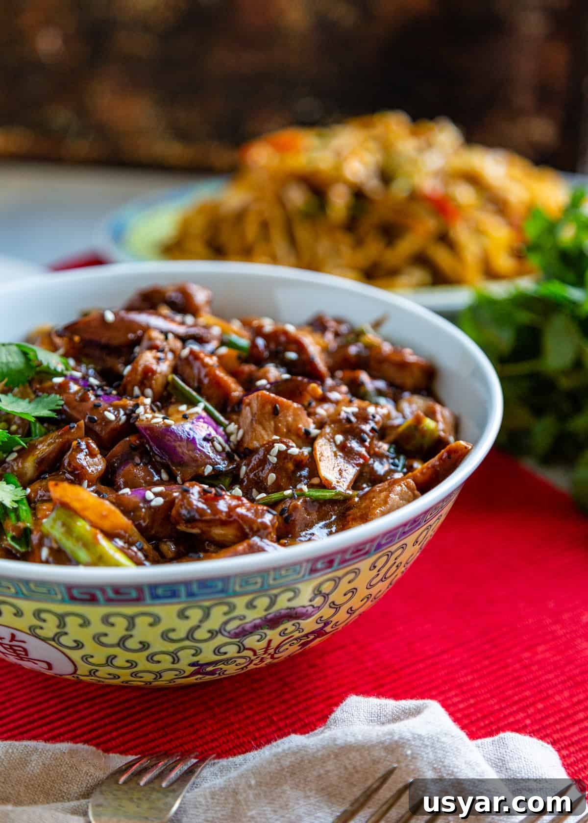 Side view: szechuan pork in serving bowl on red tablecloth with forks on white tea towel on side