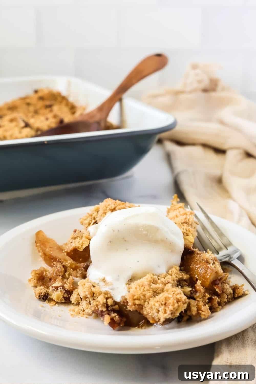 Warm Fresh Pear Crisp Recipe with Cinnamon Oat Topping 9 A serving of pear crisp made with fresh pears on a white plate, topped with vanilla ice cream. The pan of remaining crisp is in the background.