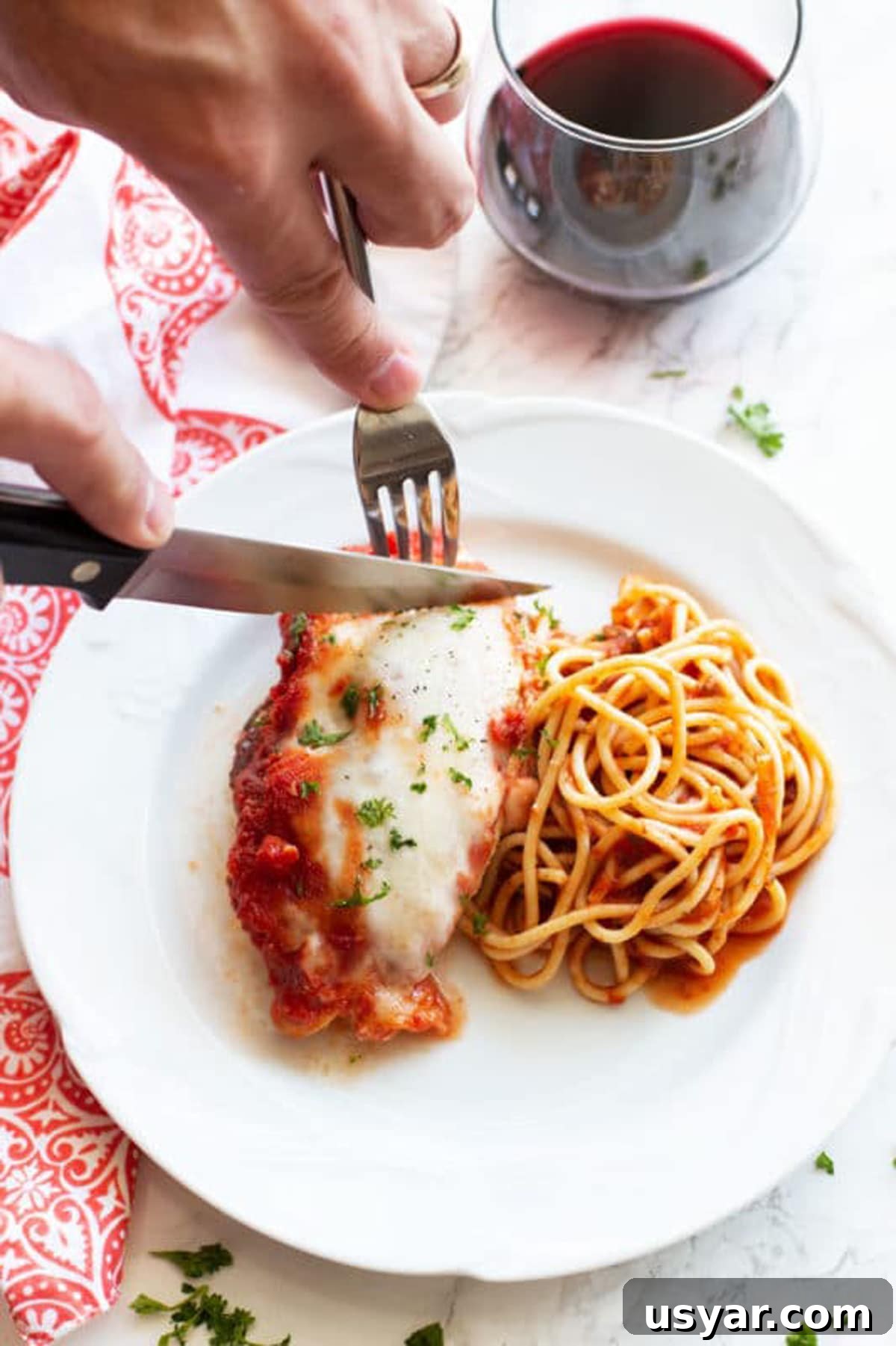 Baked Chicken Caprese with Mozzarella and Tomatoes 11 Knife cutting into a chicken breast topped with Provolone cheese served with a side of pasta. Red wine on table.