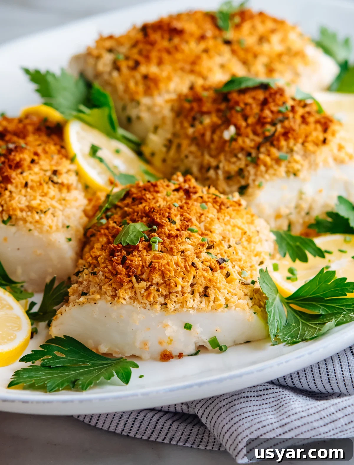 Parmesan Baked Cod with Garlic and Lemon 2 parmesan baked cod served with parsley and lemon slices on a white oval platter.