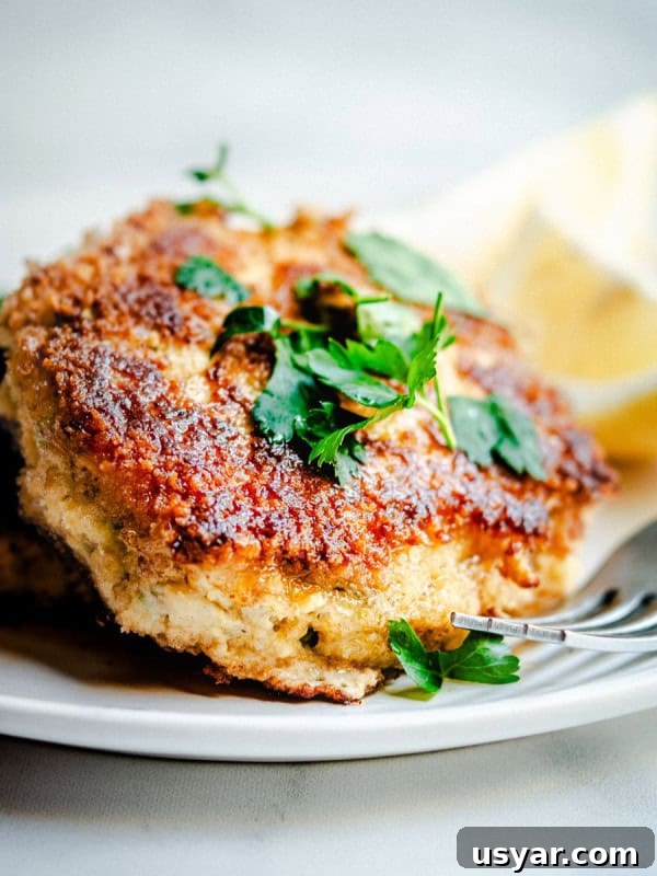 Parmesan Baked Cod with Garlic and Lemon 4 A close-up of a breaded and fried cod fish cake garnished with fresh parsley, served on a white plate. A lemon wedge can be seen in the background. A fork partially rests on the plate near the cod fish cake.