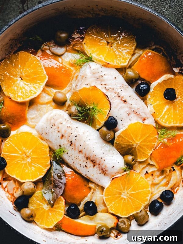 Parmesan Baked Cod with Garlic and Lemon 5 A skillet with cooked halibut fish fillets surrounded by sliced oranges, black olives, green olives, herbs, and fennel. The dish is colorful and garnished with fresh green herbs.