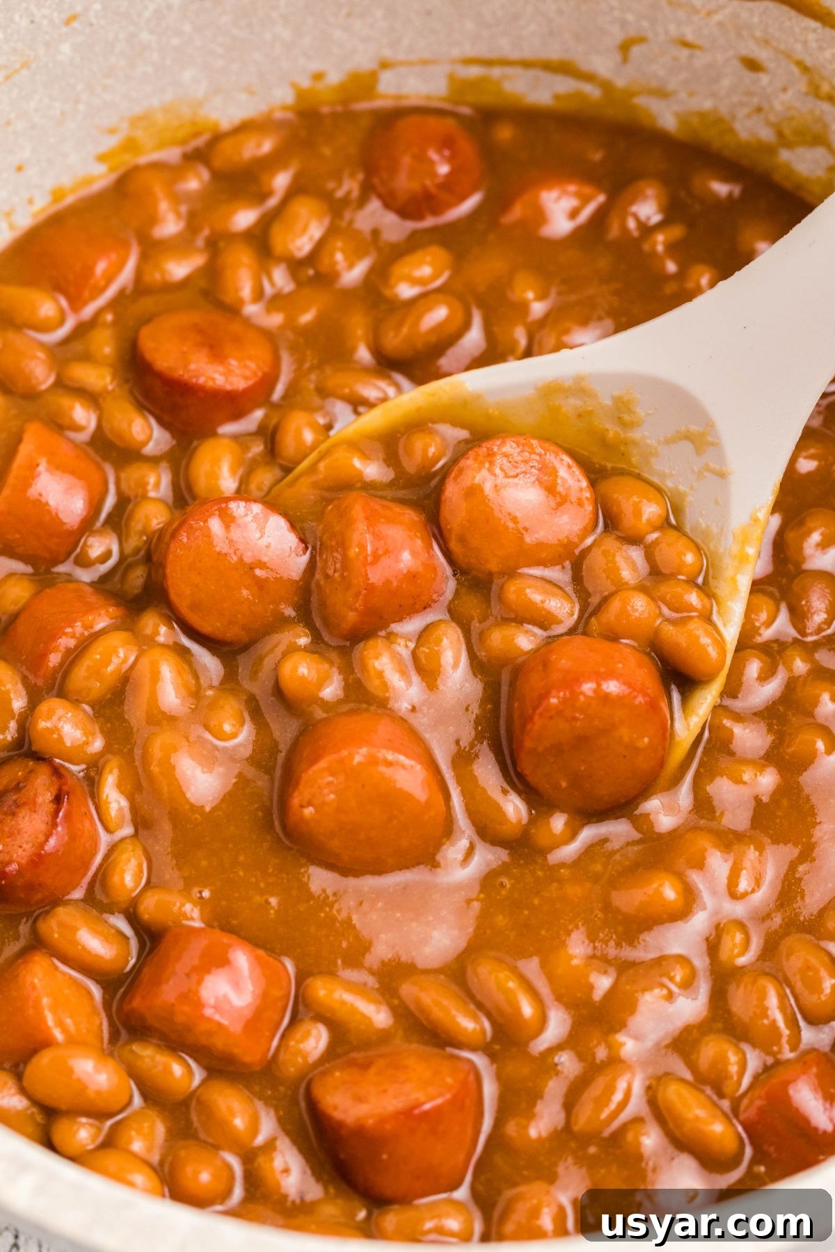 Beanie Weenies Recipe: Classic Hot Dogs in Baked Beans 7 Close up photo of beans and weenies in a saucepan with a large spoon in the center.
