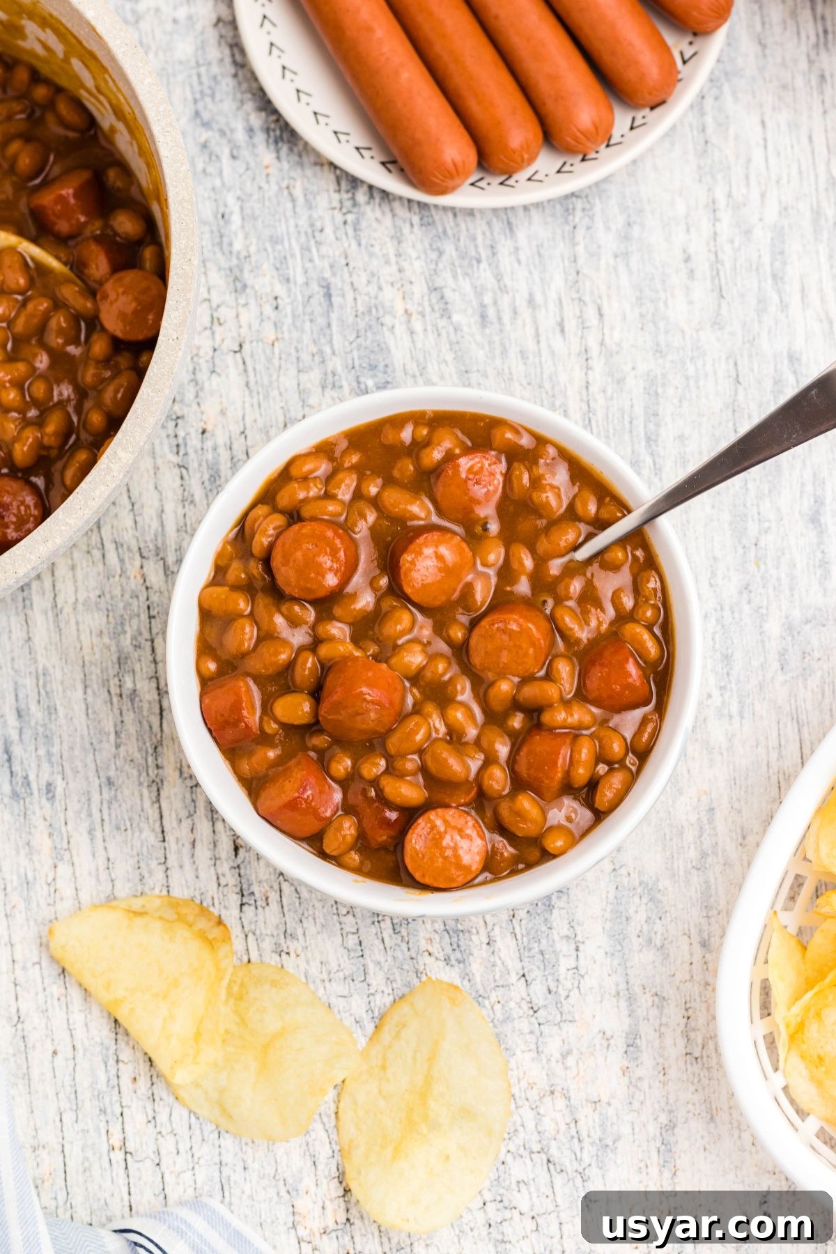 Beanie Weenies Recipe: Classic Hot Dogs in Baked Beans 8 Cut up hot dogs, mixed with beans, in a white bowl with potato chips scattered around the dish.