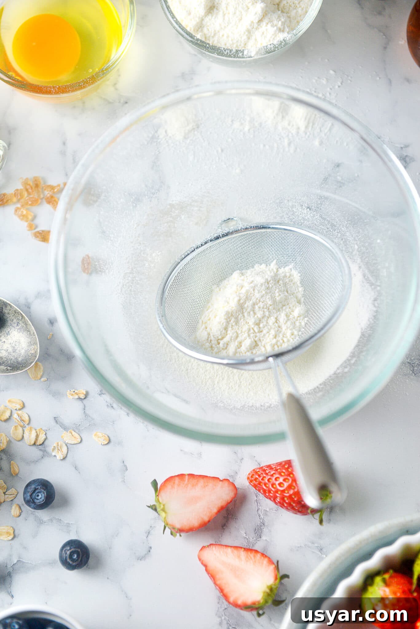 Low-Carb Dessert Baking Tips and Tricks for Perfect Results 5 Sifted dry ingredients in a clear glass bowl on a marble countertop with scattered ingredients all around.